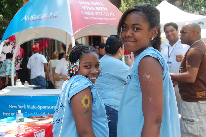 Dos mentoreadas con camisetas del programa junto al puesto de una organización de salud aliada en una feria callejera.