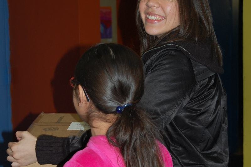 Una mujer y una niña con abrigo rosa cargan juntas una caja de cartón con donaciones.