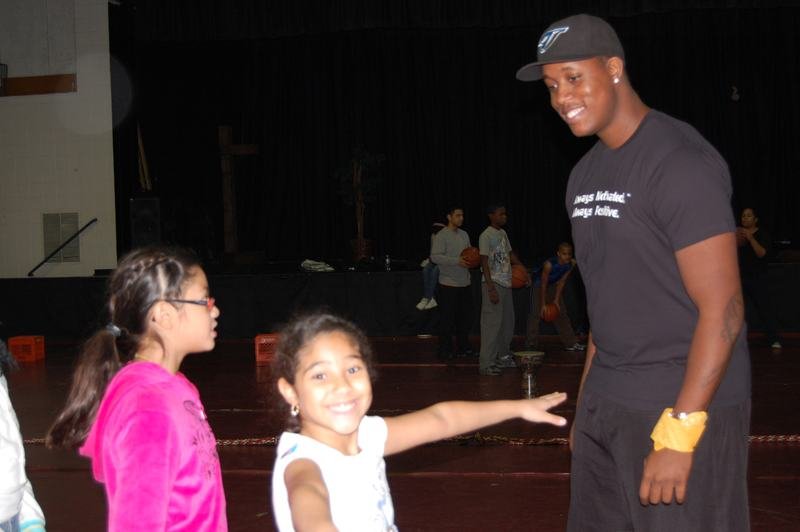 Un entrenador con gorra negra habla con dos jóvenes mentoreadas en el gimnasio poco iluminado de una escuela.