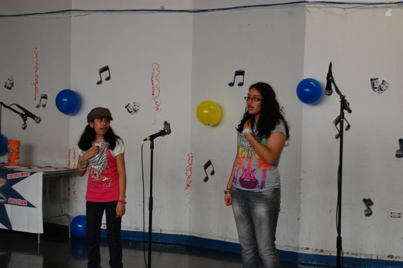 Dos mentoreadas cantan ante los micrófonos frente a una pared decorada con notas musicales de papel.